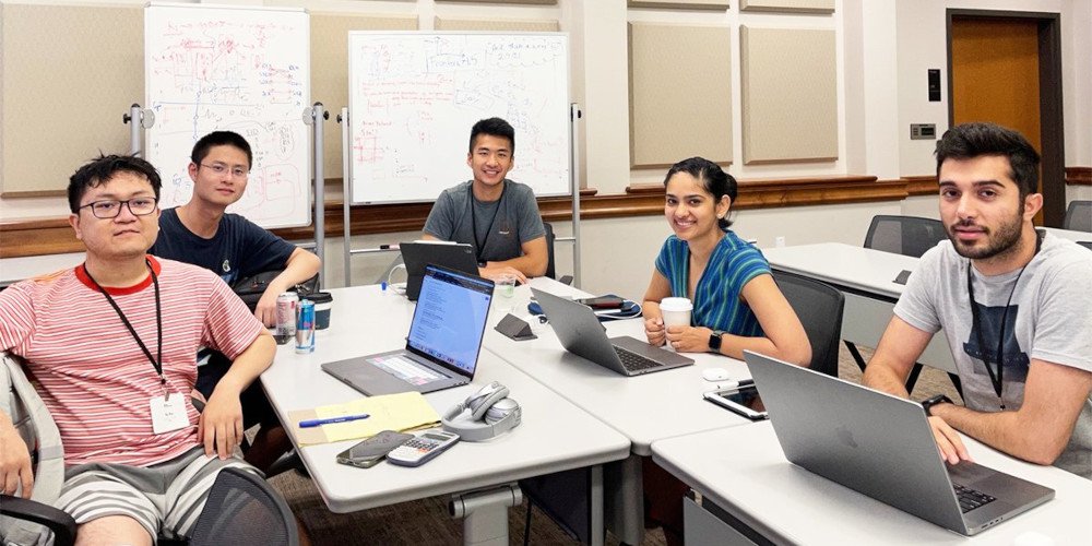 a group of people sitting around a desk with laptops